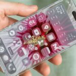 Mão segurando um iPhone com corpo transparente de vidro, mostrando ícones coloridos e um widget de clima na tela inicial.