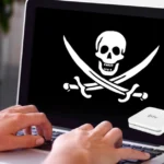 Mãos de usuário digitando em notebook com símbolo de pirataria (caveira) na tela e imagem ilustrativa de aparelho BTV Box e controle remoto no canto.