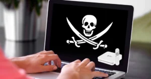 Mãos de usuário digitando em notebook com símbolo de pirataria (caveira) na tela e imagem ilustrativa de aparelho BTV Box e controle remoto no canto.